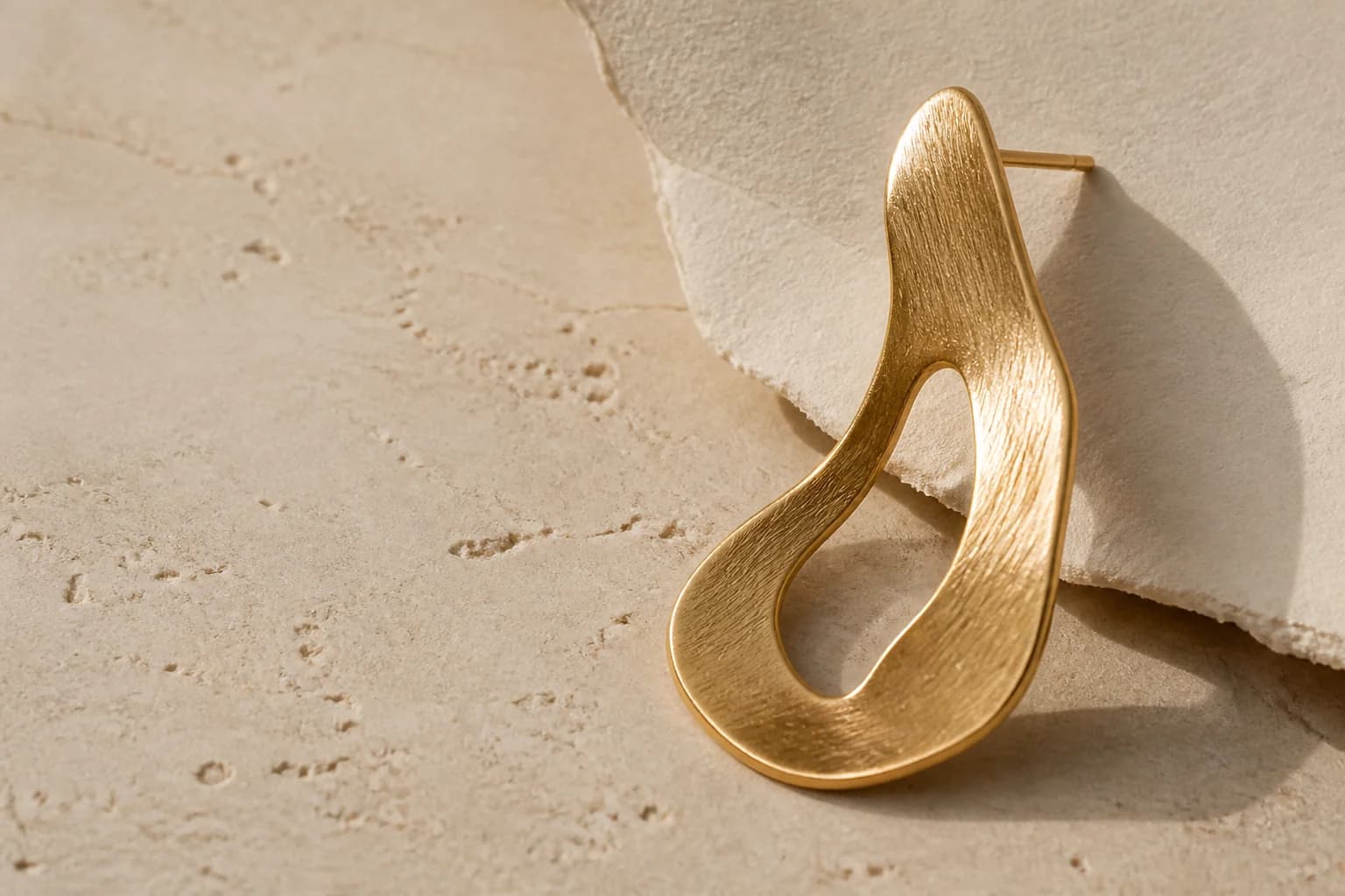 Handcrafted sculptural gold earring on warm limestone